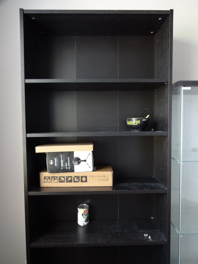 A black bookshelf that's mostly empty except for a can of beans, some small boxes, and a cracked ramen bowl.