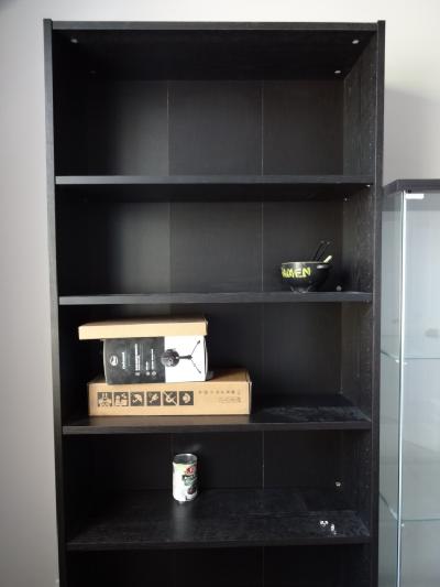 A black bookshelf that's mostly empty except for a can of beans, some small boxes, and a cracked ramen bowl.